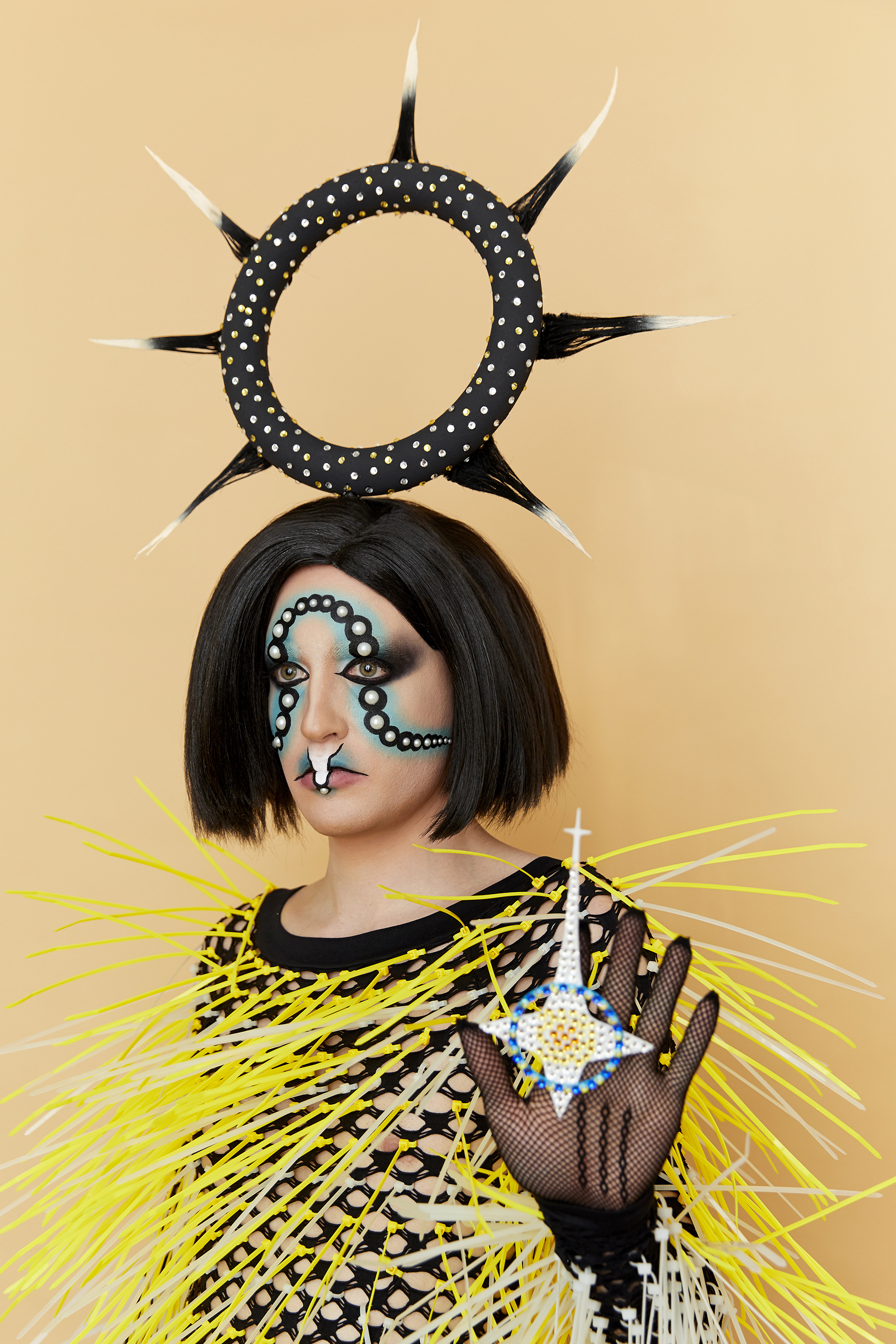 Jesse facing away from the camera wearing a graphic makeup look with an ornate headpiece and black fishnet top covered in yellow and white spikes against a tan background